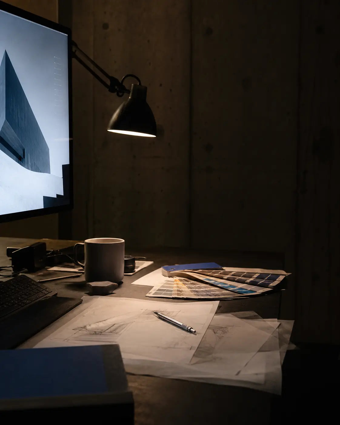 A dark design workspace with a monitor, paper sketches, color cards, pencil and coffee cup