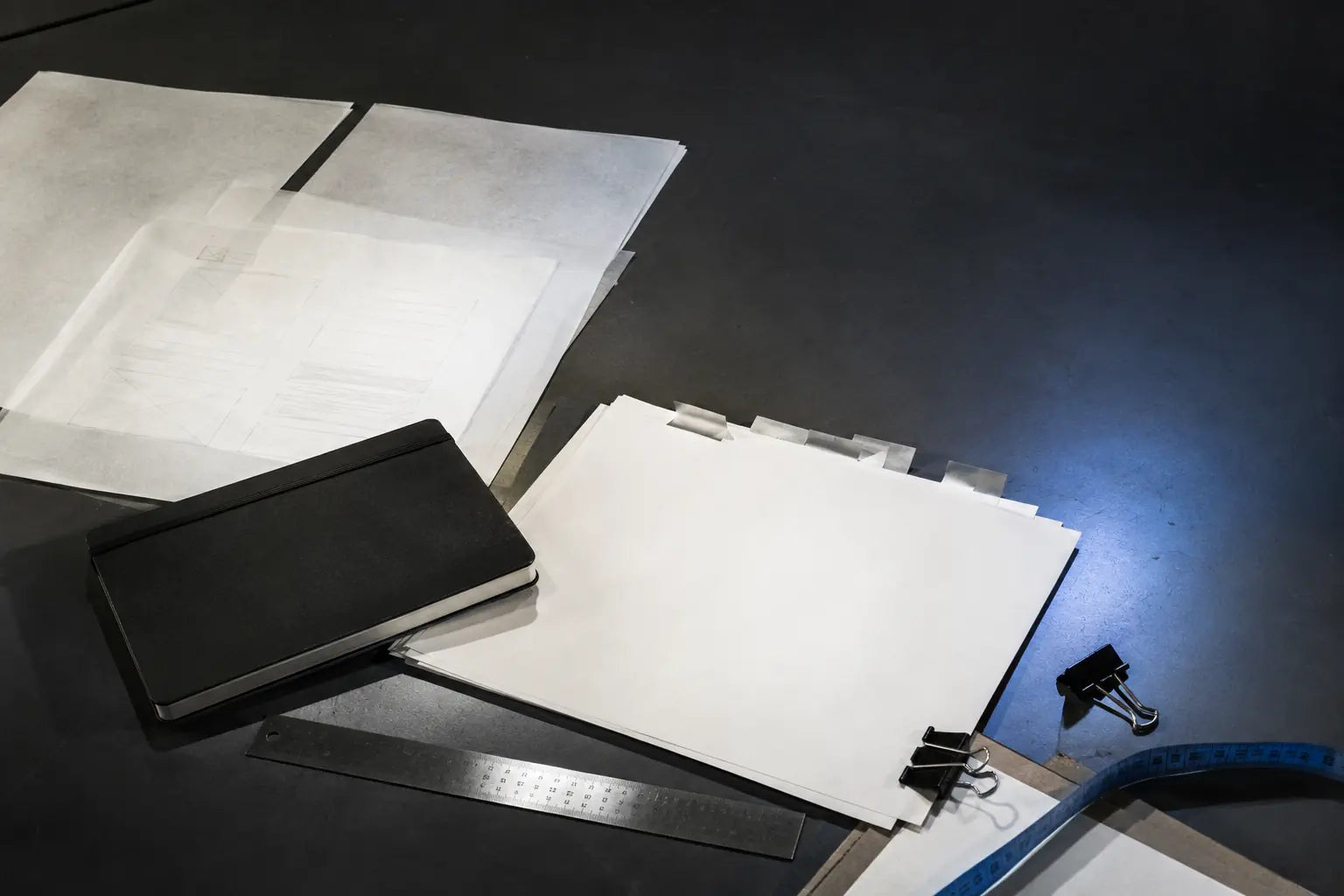 Blank project briefs, tracing-paper wireframes, a black notebook, clips, ruler, and blue measuring tape on a studio desk