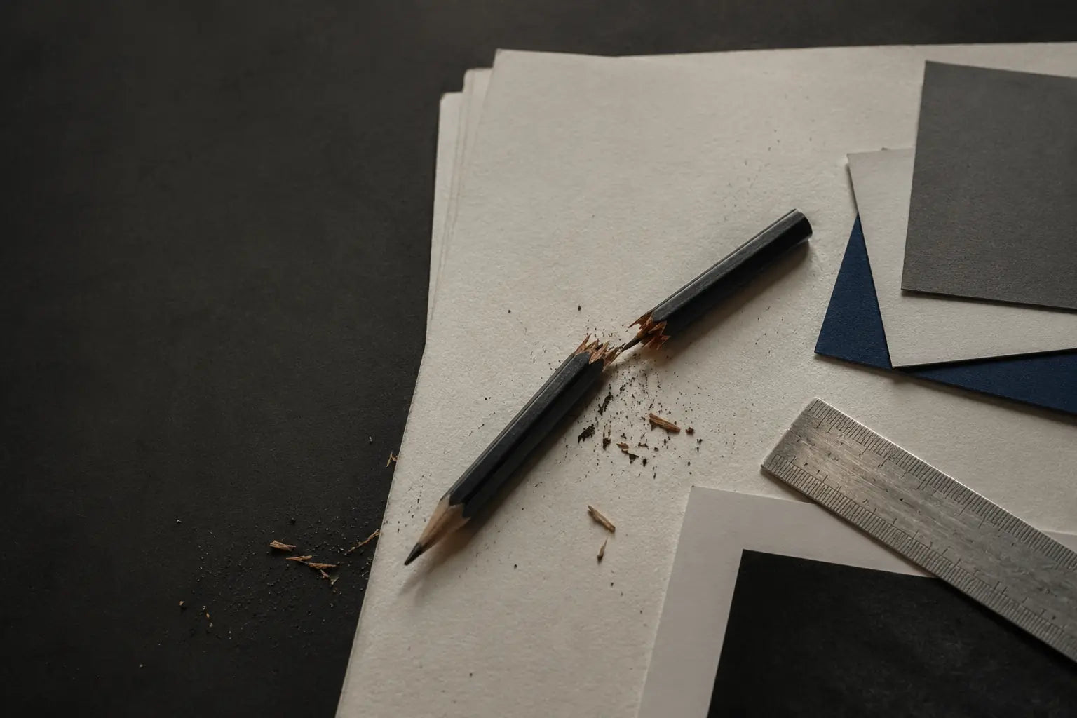 A snapped graphite pencil, paper fibers, color samples, and a metal ruler photographed in a studio