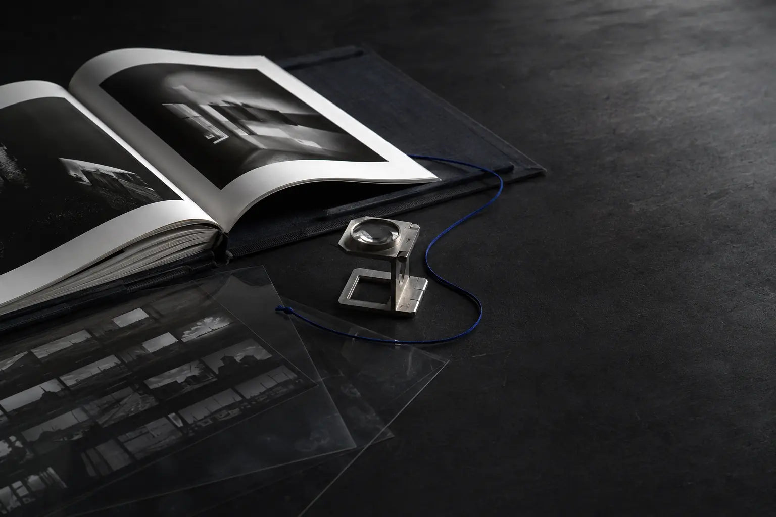 A clothbound portfolio book, photographic contact prints, and a loupe on a dark studio table