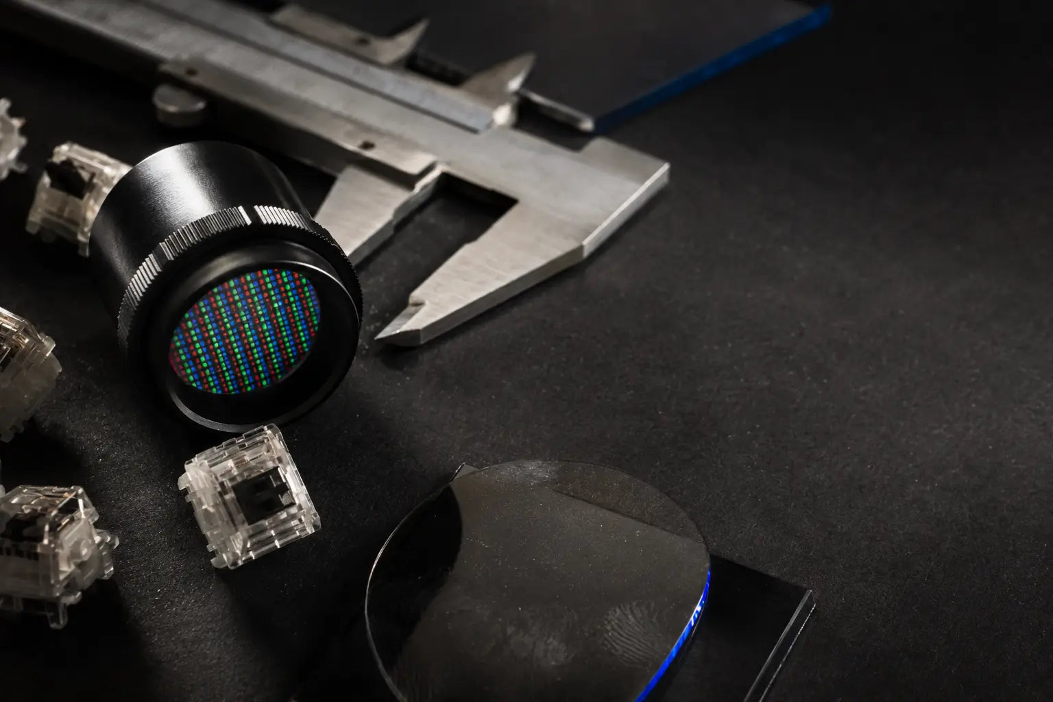 A monitor-pixel loupe, metal caliper, optical glass, and translucent keyboard switches on a workbench