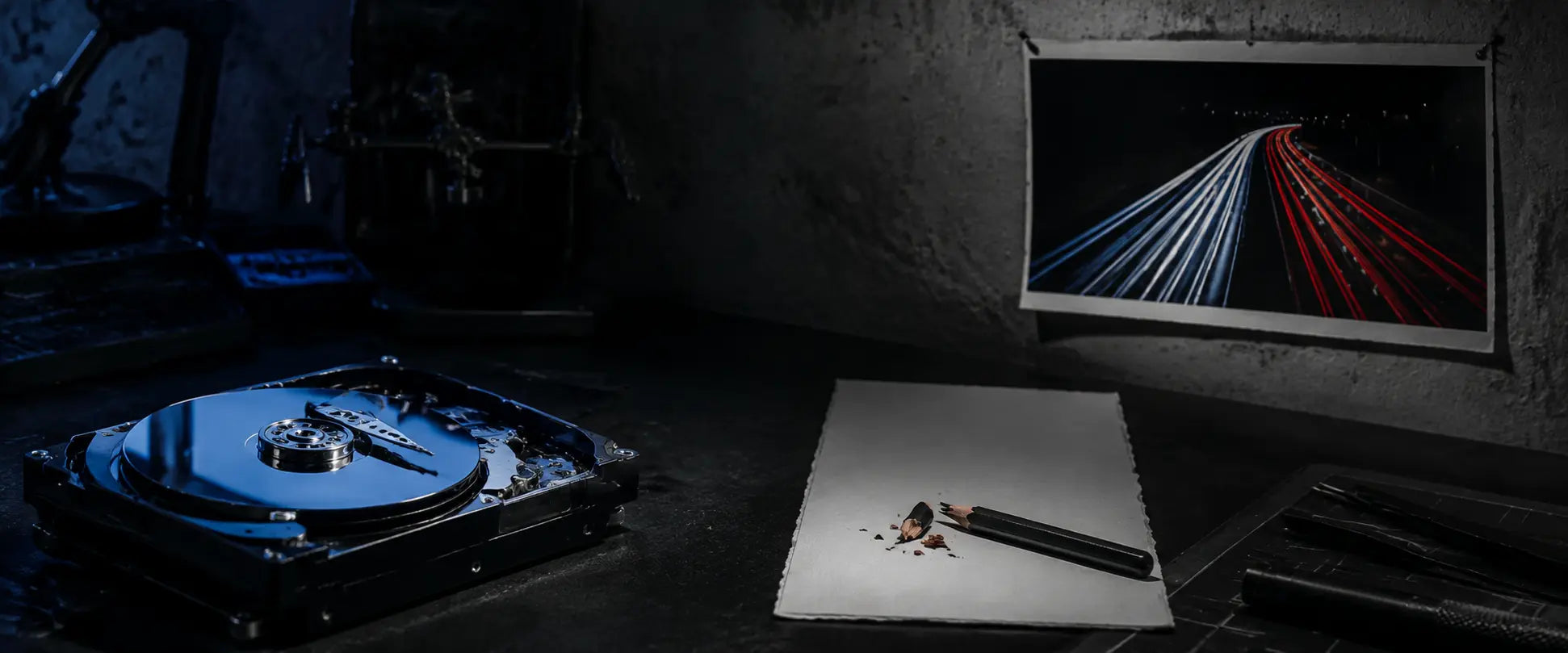 Exposed hard-drive platters, a snapped graphite pencil, and a pinned long-exposure light photograph on a studio workbench